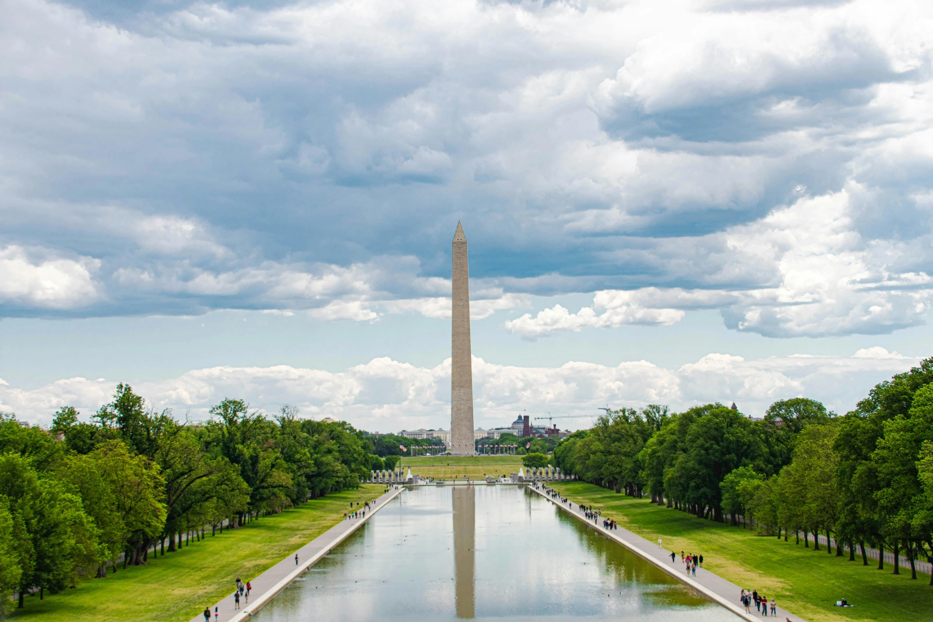 The Washington Monument over the reflecting pool at the National Mall, with several groups of people walking alongside the pool