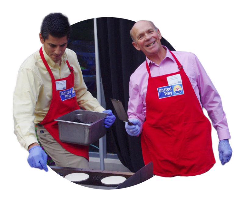 Two men in United Way aprons flipping pancakes on griddle, with their heads and arms extending outside the circle frame