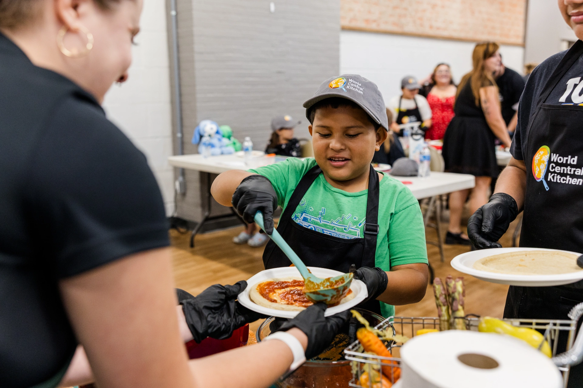 Young child helping spread pizza sauce on a personal pizza dough at a World Central Kitchen event