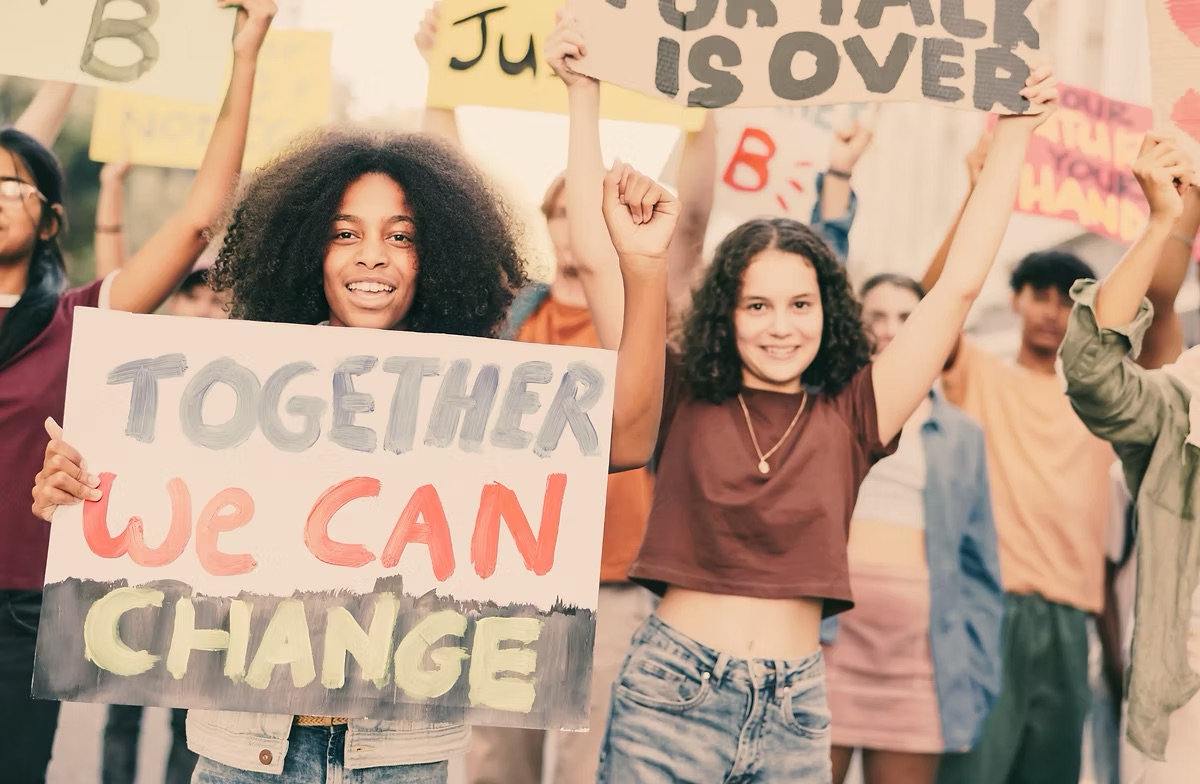 Photo of two young people, women, in a march, holding a sign that says Together We Can Change.